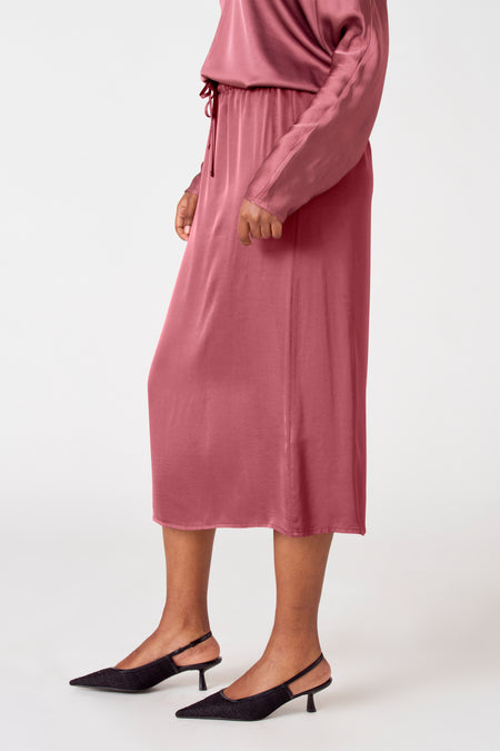 Robe rose d'Astrid Black Label, matériau soyeux en longueur midi, portée avec des sandales à talons compensés noires.