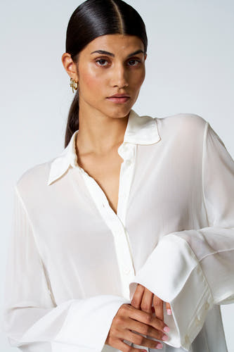 Ecru blouse met lange mouwen van Astrid Black Label, met lange manchetten en gouden oorbellen.
