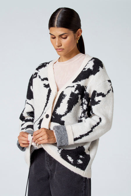 Ecru gilet van Astrid Black Label, met zwart dessin en lange mouwen, gedragen over een roze top en zwarte broek.
