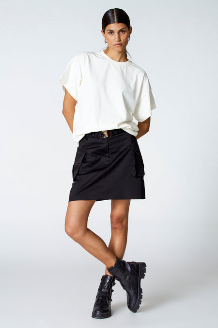 Jupe courte noire d'Astrid Black Label, avec des poches brodées, associée à un T-shirt blanc et à des bottes noires.
