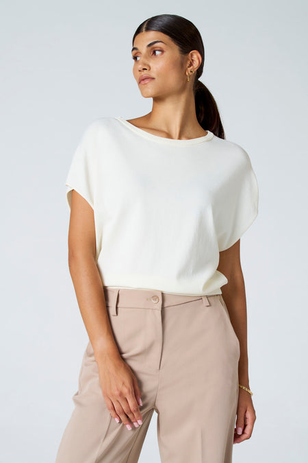 T-shirt écru d'Astrid Black Label, porté avec un pantalon beige et un bracelet en or, sur un fond blanc.