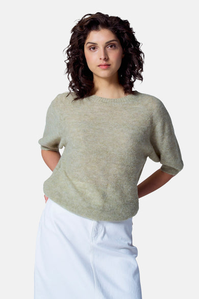 Pull vert avec col rond d'Astrid Black Label, à manches courtes, porté avec un pantalon blanc.