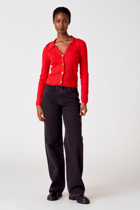 Gilet rouge d'Astrid Black Label avec des manches longues et des boutons dorés, associé à un jean noir et des chaussures noires.