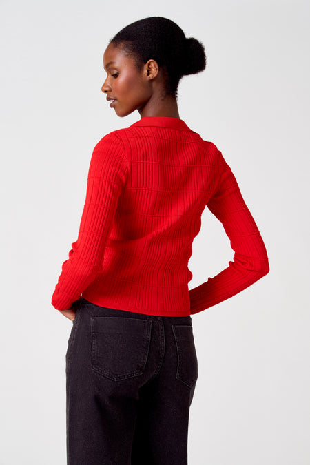 Gilet rouge d'Astrid Black Label, avec une fine structure en côtes, porté avec un jean foncé.