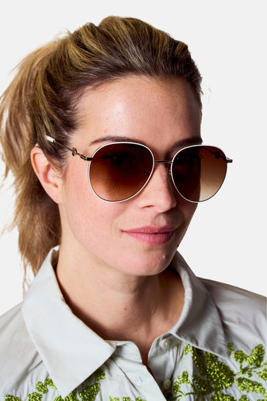 Witte zonnebril van Astrid Black Label, met een goudkleurig montuur en bruine glazen, gedragen door een vrouw met een lichtgroene blouse.
