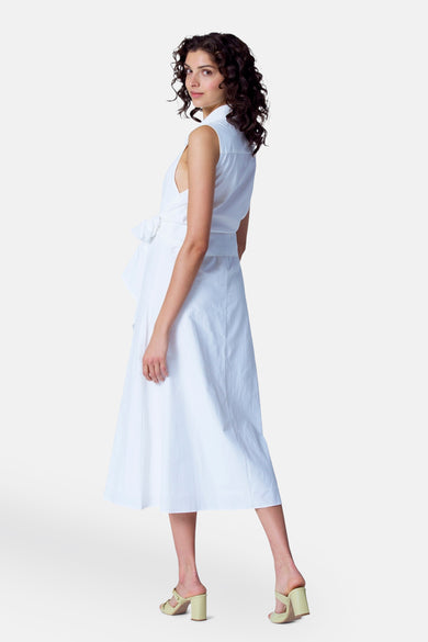 Mouwloze witte jurk van Astrid Black Label, met strikceintuur en halverwege de kuit, gedragen met lichtgele hakken.
