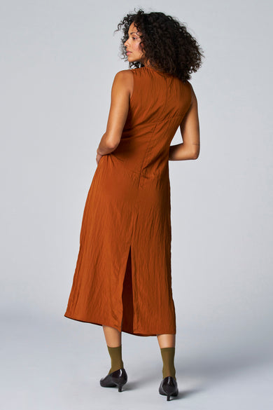 Bruine mouwloze jurk van Astrid Black Label, met een lange rok met split en groene sokken met donkerbruine hakken.
