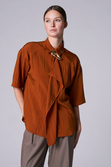Bruine blouse van Astrid Black Label met korte mouwen, geknoopte halslijn met gouden accessoire en taupekleurige broek.
