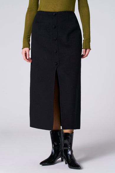 Zwarte rok van Astrid Black Label met knoopdetail en een split, gecombineerd met zwarte laarsjes en een olijfgroene top.
