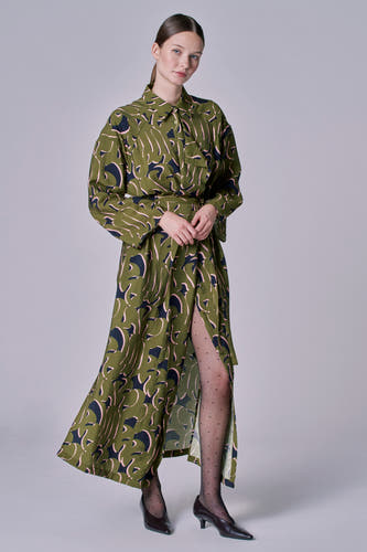 Groene jurk met lange mouwen van Astrid Black Label, met abstract bladmotief, split aan de voorkant en zwarte panty.
