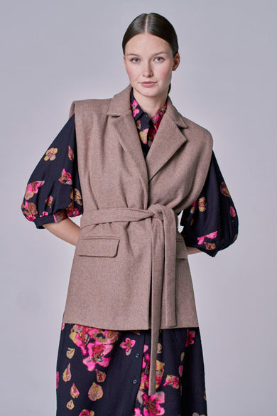 Taupekleurige blazer van Astrid Black Label, met een strikceintuur en een bloemrijke blouse en jurk eronder.
