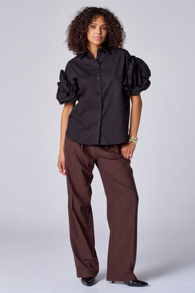 Zwarte blouse met korte mouwen van Astrid Black Label, met gepofte mouwen en gedragen met bruine pantalon en gouden armband.
