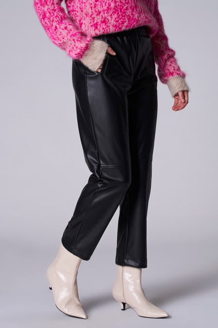 Zwarte broek van Astrid Black Label, met een leren look en gecombineerd met crèmekleurige laarsjes en een roze trui.

