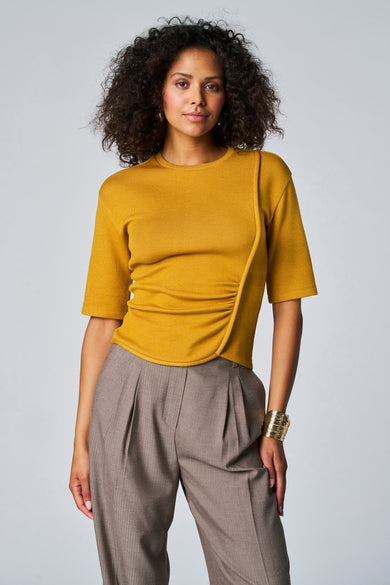 T-shirt jaune à manches courtes d'Astrid Black Label, avec des détails pliés, associé à un pantalon gris et un bracelet doré.