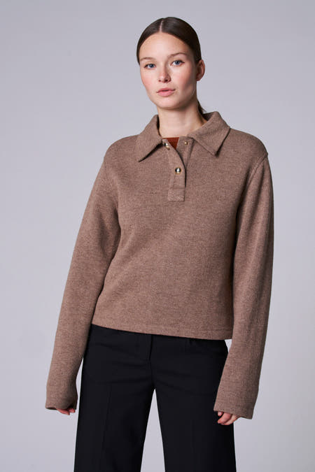Taupekleurige trui met V-hals van Astrid Black Label, met lange mouwen, gedragen met een zwarte broek.
