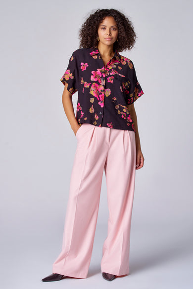 Pantalon de couleur rose d'Astrid Black Label, avec des jambes larges et une ceinture pliée, associé à une blouse imprimée de fleurs.