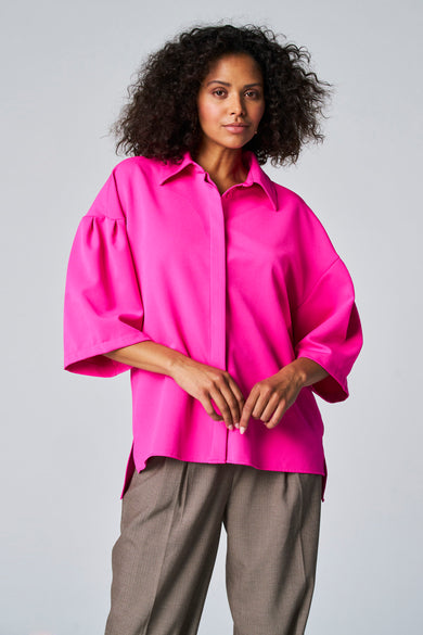 Blouse rose d'Astrid Black Label, avec des manches trois-quarts et portée avec un pantalon marron.
