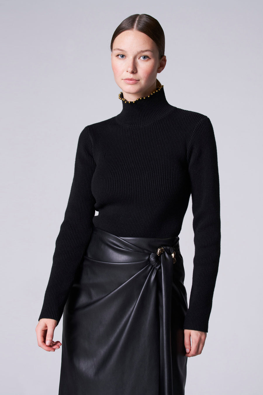 Pull-over noir à col roulé d'Astrid Black Label, avec bordure en perles d'or et une jupe en cuir noire avec détail de nœud.