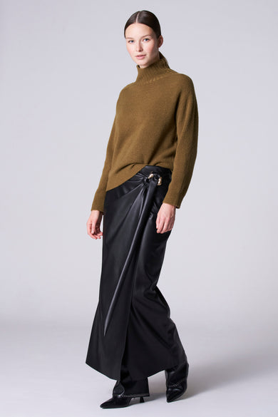 Groene trui met rolkraag van Astrid Black Label, gedragen met een zwarte leren rok met wikkeldetail en zwarte schoenen.
