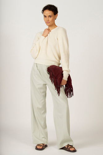 Een vrouw met een gebroken witte trui, lichtgroene broek, en een bordeauxrode tas met franjes.