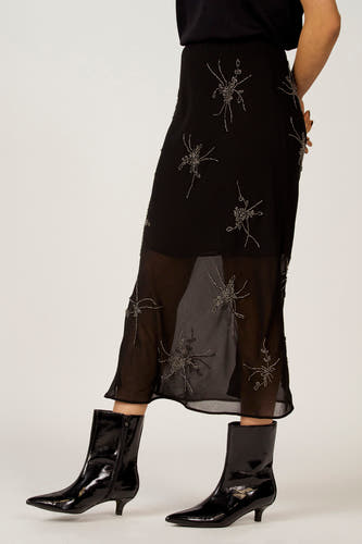 Zwarte rok van Astrid Black Label met zilverkleurige sierdetails en een transparante zoom, gecombineerd met zwarte, glanzende enkellaarsjes.

