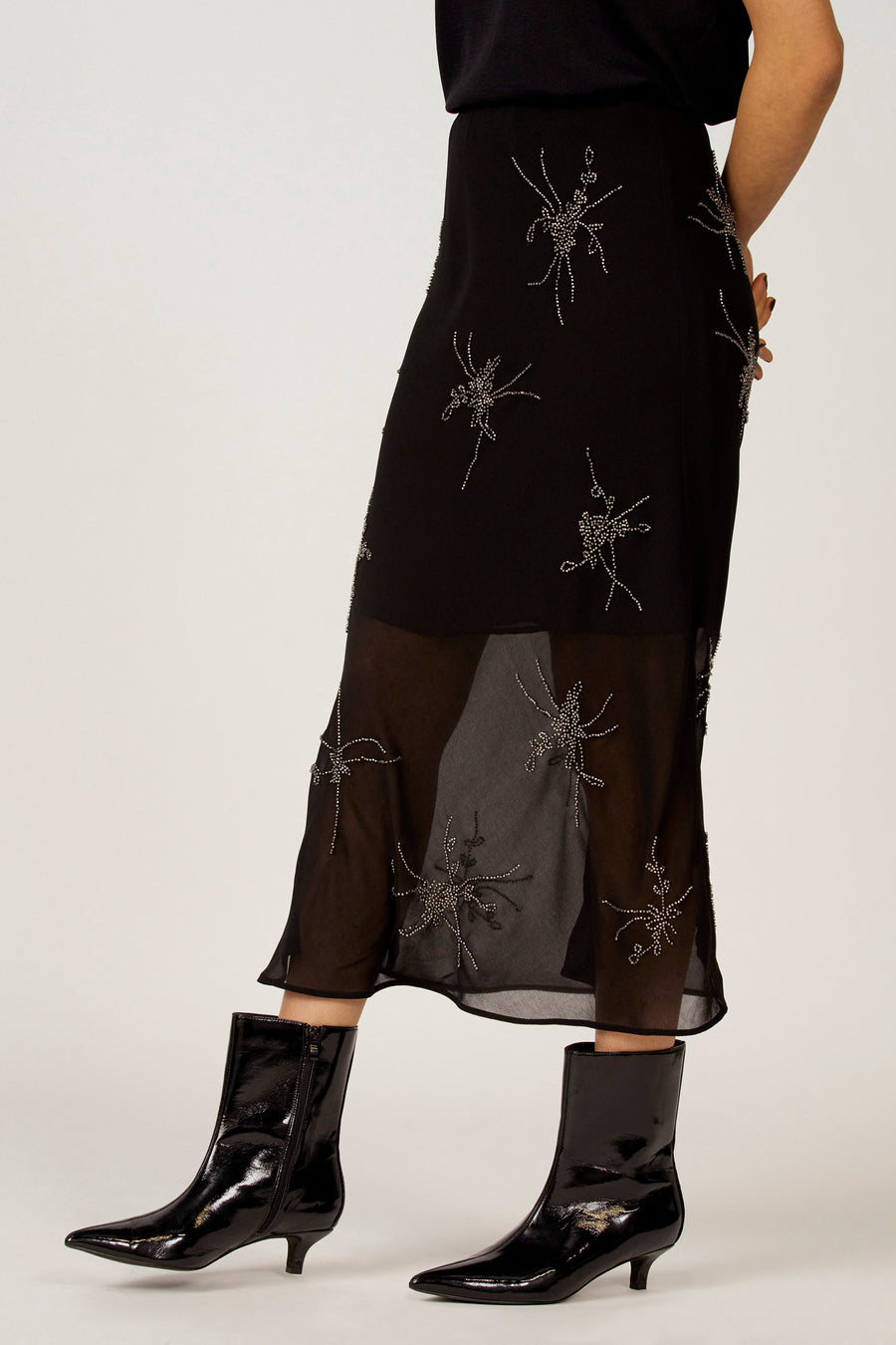 Zwarte rok van Astrid Black Label met zilverkleurige sierdetails en een transparante zoom, gecombineerd met zwarte, glanzende enkellaarsjes.
