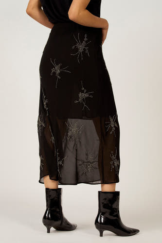 Zwarte rok van Astrid Black Label met zilveren decoratieve bloemmotieven en transparante zoom, gezien van achteren, gecombineerd met zwarte laarsjes.
