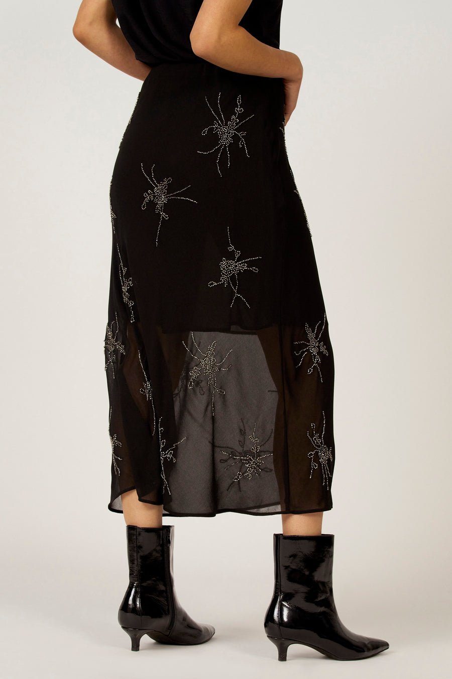 Zwarte rok van Astrid Black Label met zilveren decoratieve bloemmotieven en transparante zoom, gezien van achteren, gecombineerd met zwarte laarsjes.
