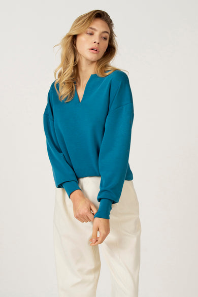 Pull-over bleu à col V d'Astrid Black Label, coupé loose et associé à un pantalon crème.