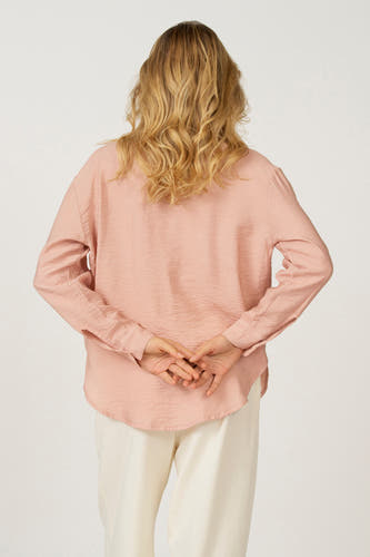Blouse rose à longues manches d'Astrid Black Label, avec les mains croisées sur le dos et un short blanc.