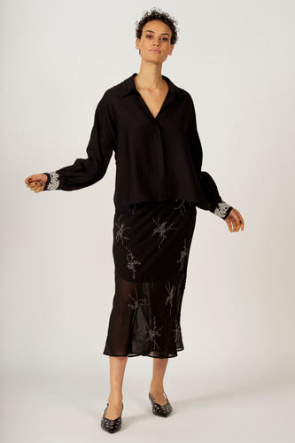 Chemise noire à longues manches d'Astrid Black Label, avec des détails argentés sur les poignets. Portée avec une jupe et des chaussures.