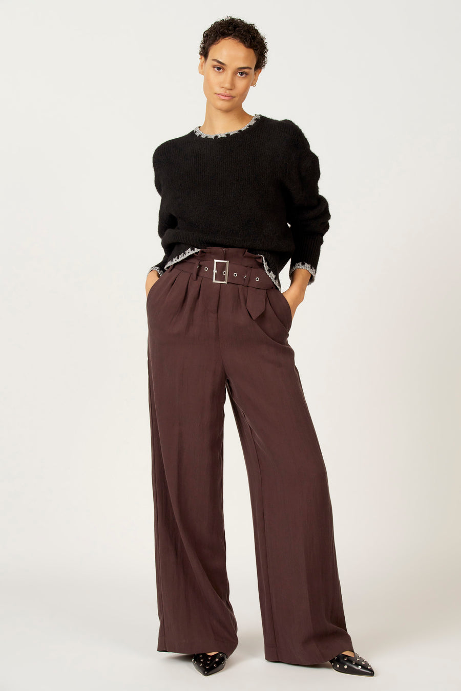 Vrouw met zwarte trui en bruine broek met riem, staand tegen witte achtergrond.