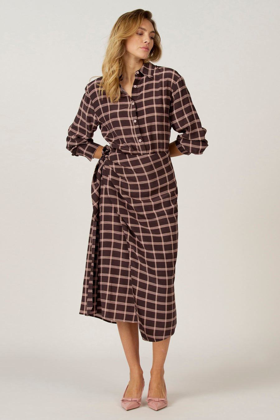 Bruine jurk met lange mouwen van Astrid Black Label, met een geruite patroon, gedragen met roze schoenen met strik.
