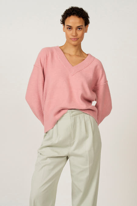 Roze V-hals trui van Astrid Black Label, gedragen met een lichtgroene pantalon en gouden oorbellen.
