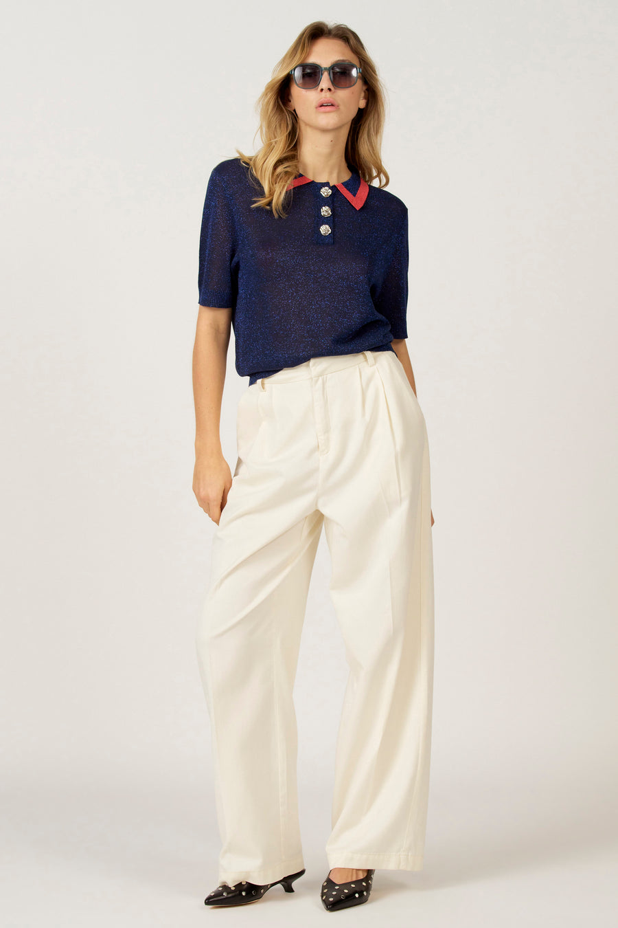 Vrouw in blauwe top met rode kraag, witte broek en zwarte schoenen.