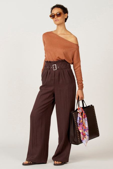 Vrouw in koperkleurige top en bruine pantalon met riem, draagt een zwarte tas met sjaal en sandalen, tegen een witte achtergrond.