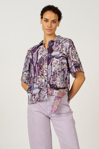 Chemisier multicolore à manches courtes d'Astrid Black Label, avec un imprimé abstrait violet et porté avec un pantalon lilas.