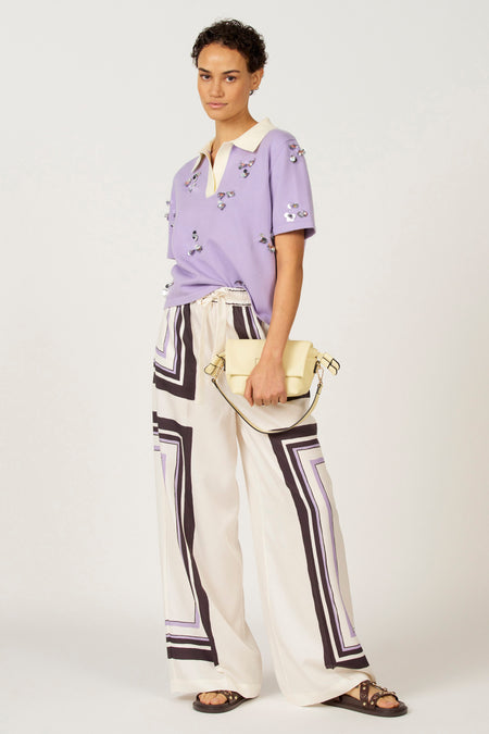 Paarse polo met korte mouwen van Astrid Black Label, versierd met zilveren pailletten, gecombineerd met een witte broek met geometrisch patroon en sandalen.
