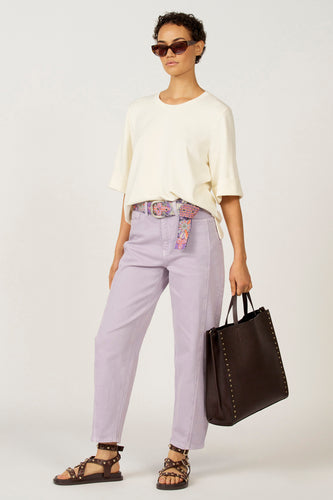 Vrouw in lila broek, wit shirt, bloemenriem en donkerbruine tas.