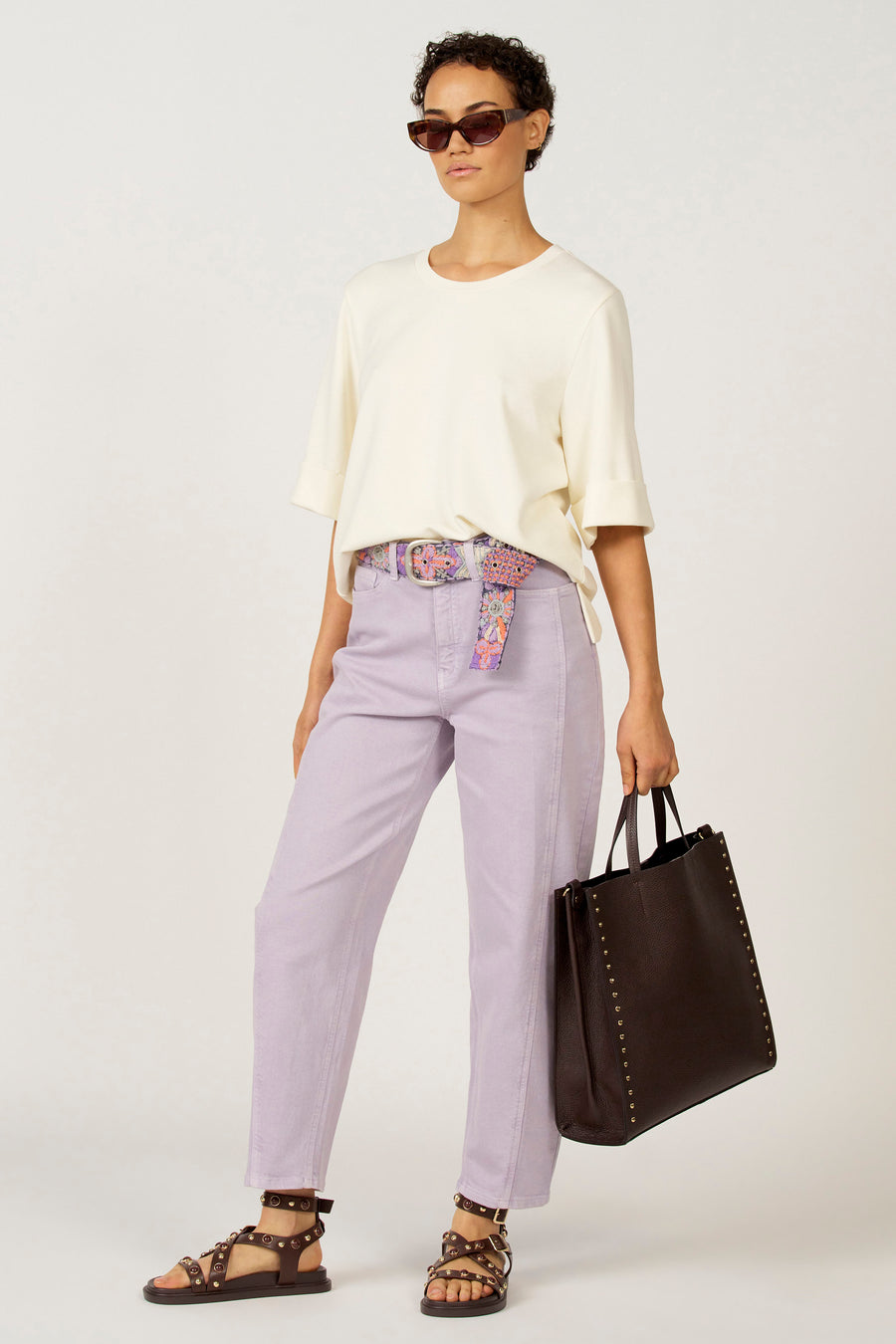 Vrouw in lila broek, wit shirt, bloemenriem en donkerbruine tas.