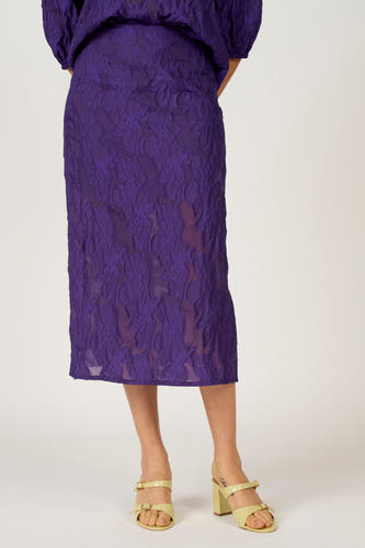 Paarse lange rok van Astrid Black Label, met een bloemmotief en textuur, gecombineerd met gele sandalen met hak.
