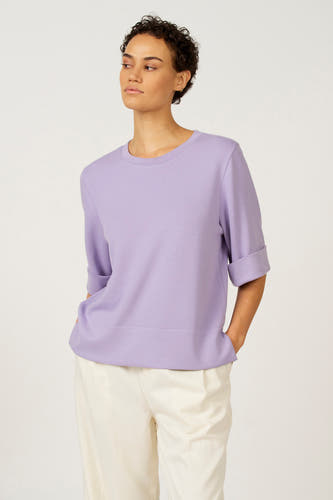 T-shirt violet à manches courtes d'Astrid Black Label, avec des manches retroussées et porté avec un pantalon blanc.