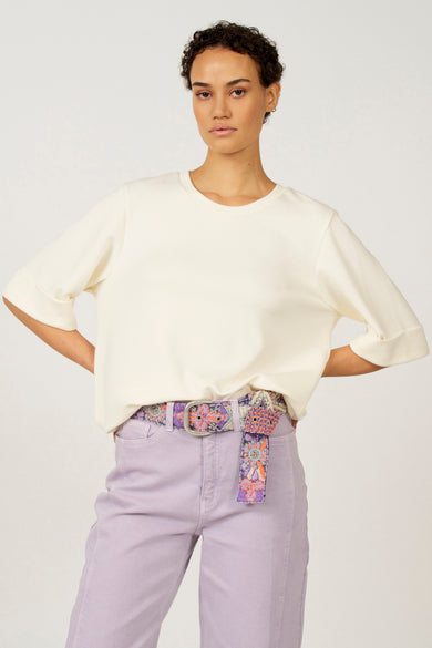 T-shirt écru à manches courtes d'Astrid Black Label, avec des manches retroussées et une fente latérale, associé à un pantalon violette.