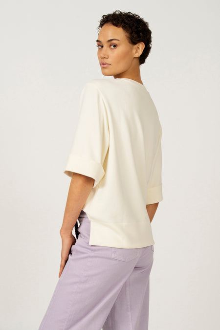 Ecru T-shirt met korte mouwen van Astrid Black Label, met omgeslagen mouwen en zijsplit, gecombineerd met een lila broek.
