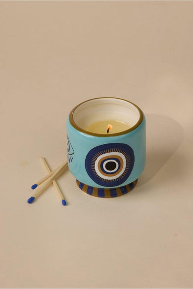 Multikleurige kaars van PADDY WAX, met decoratieve blauwe en gouden oogmotieven op een keramische houder, naast twee lucifers.
