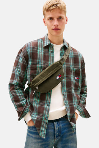 Sac à dos vert de TOMMY JEANS, porté sur une chemise rayée marron et turquoise avec un pull blanc et des jeans bleus.