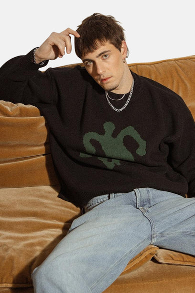 Pull noir avec col rond de KAOTIKO, imprimé vert, associé à des jeans et des chaînes d'argent.