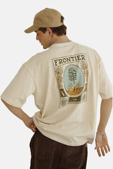 Ecru T-shirt met korte mouwen van KAOTIKO, met een print op de rug, gedragen met een bruine broek en pet.
