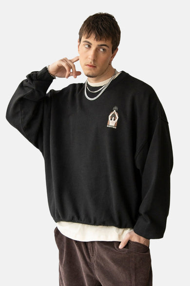Pull-over noir de KAOTIKO, avec un petit imprimé sur la poitrine et porté avec des chaînes et un pantalon marron.