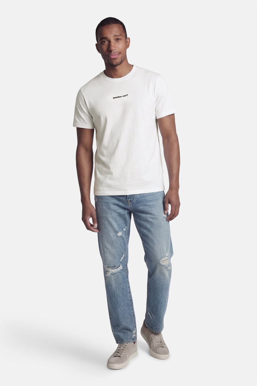 Wit T-shirt van KAOTIKO, met korte mouwen en een zwarte tekstprint, gecombineerd met blauwe ripped jeans en grijze schoenen.